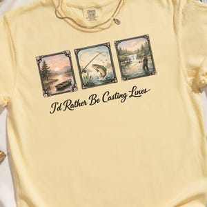 May include: Yellow t-shirt with three framed fishing scenes and the text "I'd Rather Be Casting Lines" in black script. The scenes depict a boat on a lake, a fish jumping, and a person fishing near a waterfall. A necklace is visible at the neckline.