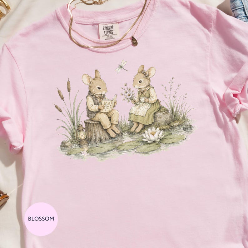 Pink Comfort Colors shirt with a print of two rabbits reading and holding flowers by water.
