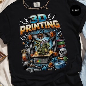 May include: Black t-shirt with a colorful graphic featuring a 3D printer, a green dragon, and the words "3D PRINTING" in a stylized font. The design includes spools of filament, a skull, and various mechanical components.