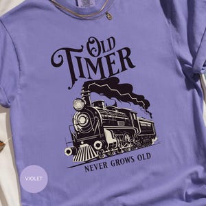 May include: A violet t-shirt with a black and white illustration of a steam train. The text "Old Timer" is above the train, with "Never Grows Old" below. The shirt has a crew neck and short sleeves.