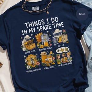 Op de afbeelding: Een marineblauw t-shirt met de tekst "THINGS I DO IN MY SPARE TIME" en zes illustraties over bijenhouden: bijenkasten controleren, honing oogsten, beschermende kleding aantrekken, de koningin beschermen, honing bottelen en praten over bijen.