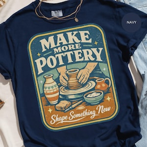 May include: Navy blue t-shirt with a retro graphic that reads "MAKE MORE POTTERY" above an illustration of hands shaping clay on a pottery wheel. The design includes pottery tools and finished pieces. The text "Shape Something New" is below the illustration.