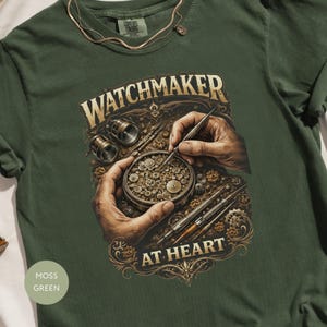 Könnte beinhalten: Moosgrünes T-Shirt mit detaillierter Grafik. Die Grafik zeigt Hände, die mit Werkzeugen an Uhrwerken arbeiten, mit dem Text "WATCHMAKER AT HEART". Das T-Shirt hat einen Rundhalsausschnitt und kurze Ärmel.