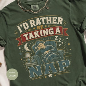 May include: A moss green t-shirt featuring a graphic print of a person sleeping in an armchair under a blanket. The text reads I'D RATHER BE TAKING A NAP. The design includes a crescent moon, a lamp, and stars in shades of cream, red, and blue.