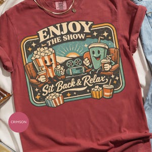 May include: Crimson-colored t-shirt with a retro movie-themed graphic. The design features cartoon popcorn and a drink enjoying a movie, with the text "ENJOY THE SHOW - Sit Back & Relax". The shirt has a crew neck and short sleeves.