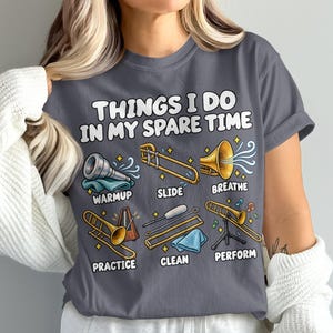Könnte beinhalten: Ein dunkelgraues T-Shirt mit dem Text "THINGS I DO IN MY SPARE TIME" über Illustrationen von Posaunen-Aktivitäten, beschriftet mit "WARMUP", "SLIDE", "BREATHE", "PRACTICE", "CLEAN" und "PERFORM" sowie verschiedenen Musikinstrumenten.