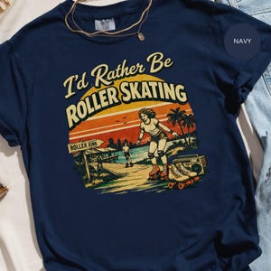 Puede incluir: Camiseta azul marino con un gráfico retro que dice "I'd Rather Be Roller Skating". El diseño presenta una patinadora, palmeras, una puesta de sol y un radiocasete. La camiseta es de la marca Comfort Colors.