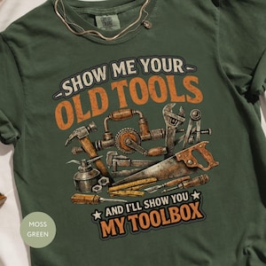 May include: Moss green t-shirt with the text "SHOW ME YOUR OLD TOOLS AND I'LL SHOW YOU MY TOOLBOX" in orange and white. The graphic features various vintage tools. A gold necklace is draped on the shirt.