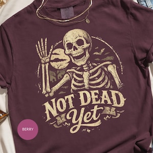 May include: Berry-colored t-shirt featuring a skeleton graphic with the text "NOT DEAD Yet". The skeleton is holding a moon and is surrounded by bats. The shirt has a crew neck and short sleeves.