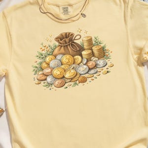 Puede incluir: Camiseta amarillo claro con un gráfico de un monedero marrón que se desborda con monedas de oro, plata y bronce. Las monedas tienen diseños de estrellas, corazones y números. La camiseta tiene el texto "COMFORT COLORS" en la etiqueta.