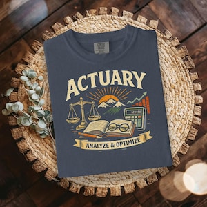 以下が含まれることがあります： ネイビーブルーのTシャツ。グラフィックデザインには、天秤、開いた本、電卓、山の風景の上に「ACTUARY」の文字が描かれています。「ANALYZE & OPTIMIZE」の文字がバナーに表示されています。Tシャツは織り込まれた表面に表示されています。