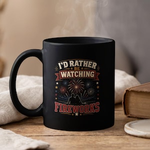 Peut inclure: Un mug en céramique noir avec une impression graphique portant le texte I'D RATHER BE WATCHING FIREWORKS. Le motif représente la silhouette de deux personnes regardant des feux d'artifice colorés dans un ciel sombre, avec des bannières rouges.