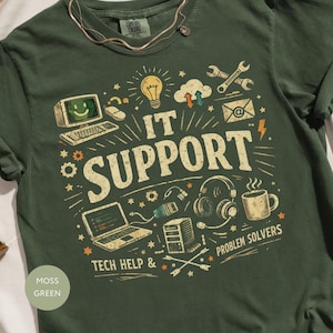 May include: Moss green t-shirt with a graphic design featuring IT support-related icons and the words "IT SUPPORT", "TECH HELP &" and "PROBLEM SOLVERS". The shirt has a crew neck and short sleeves.
