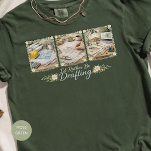May include: Moss green t-shirt with a graphic that says "I'd Rather Be Drafting." The graphic features three panels with drafting tools, blueprints, and a laptop. The shirt has a crew neck and short sleeves.