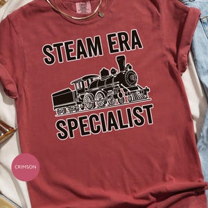 May include: Crimson-colored t-shirt with the words "STEAM ERA SPECIALIST" in white block letters above and below a black and white illustration of a steam train. The shirt is folded, with a gold necklace visible.
