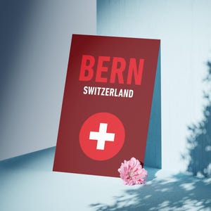May include: A maroon sign with the word "BERN" in large red letters and "SWITZERLAND" below. A white cross in a red circle is at the bottom. A pink flower rests near the sign, with shadows cast across a light blue surface.