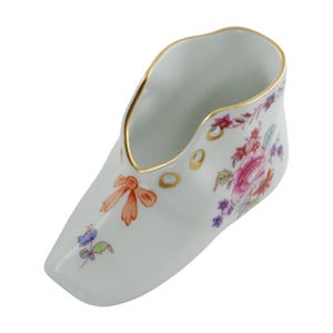 May include: A white porcelain shoe-shaped vase with a gold rim and accents. The vase is decorated with a floral pattern in pink, red, and purple, and a bow detail. The shoe has a small opening at the top.