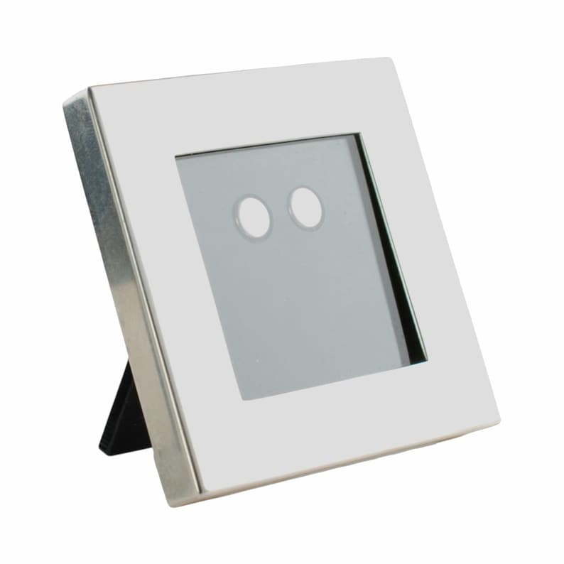 May include: A square, white picture frame with a silver metal trim. The frame has a black easel back for display. The frame's interior has a gray background with two white circles.