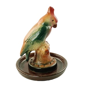 May include: A ceramic parrot figurine in shades of red, green, and tan, perched on a brown base with a circular dish. The bird has a detailed, textured appearance, with a curved beak and a prominent crest. The dish has a series of small holes.