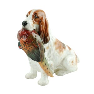 May include: A ceramic figurine of a white and brown spaniel dog sitting in an upright position. The dog has long, floppy ears and is holding a colorful game bird, featuring shades of red, brown, and green, firmly in its mouth.