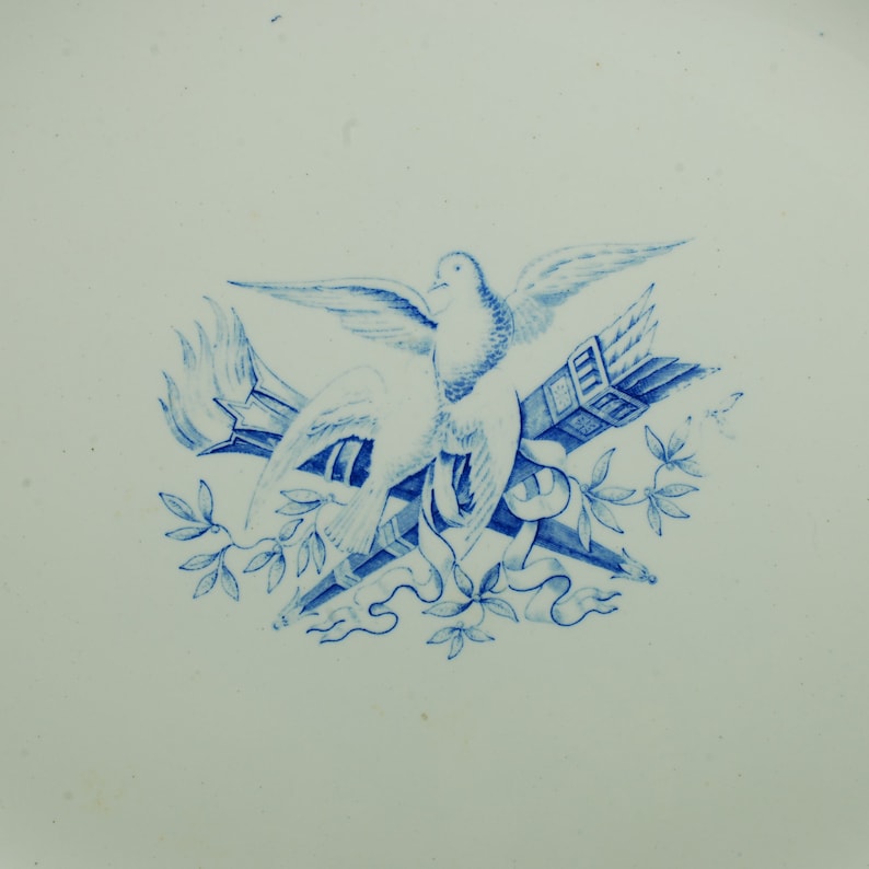 May include: Blue and white transferware design of a dove with wings spread, holding a torch and a book, surrounded by leaves.