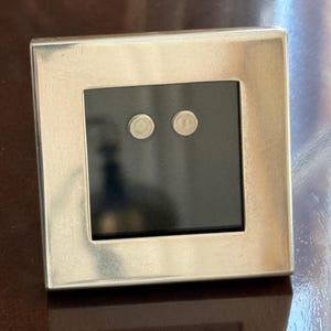 May include: A square, silver-toned picture frame with a black interior. Two small, round, silver-colored objects are centered within the black space. The frame is sitting on a dark brown surface.
