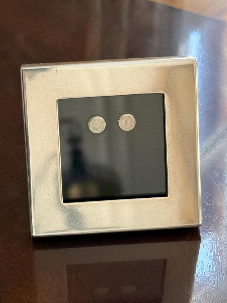 May include: A square, silver-toned picture frame with a black interior. Two small, round, silver-colored objects are centered within the black space. The frame is sitting on a dark brown surface.