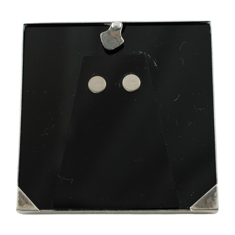 May include: Square silver-toned picture frame with a black backing. The frame has a simple, elegant design with silver corner accents and a metal clasp at the top. Two small, round magnets are visible on the back.