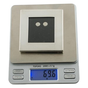 May include: A silver-toned square object with a black center and two small, round, silver-toned accents is placed on a digital scale. The scale displays a weight of 69.6 grams.
