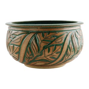 May include: A round ceramic planter with a wide opening and a detailed leaf pattern. The planter has a brown base with green accents in the carved leaf design and a green rim.