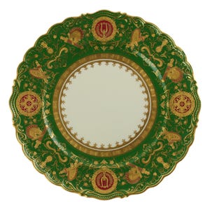 May include: A decorative green plate with a scalloped edge. The plate features gold and red accents, including ornate designs and a central cream-colored circle. The plate's rim is detailed with gold trim.