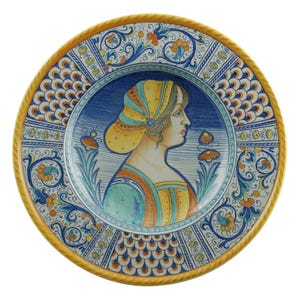 May include: A ceramic plate with a portrait of a woman in profile, wearing a yellow headscarf and gold necklace. The plate has a blue and white floral border with orange accents and a yellow rope-like edge.