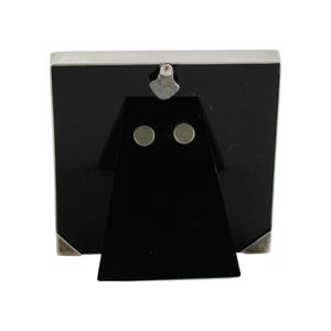 May include: A silver-toned metal picture frame with a black backing and a black easel stand. The frame has a rectangular shape with silver corner accents and a silver top. The easel stand has two circular magnets.