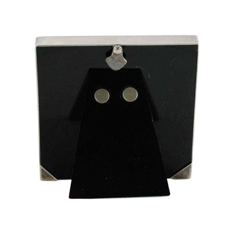 May include: A silver-toned metal picture frame with a black backing and a black easel stand. The frame has a rectangular shape with silver corner accents and a silver top. The easel stand has two circular magnets.