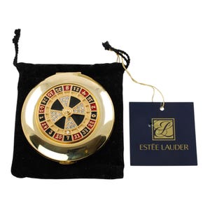 Peut inclure: Un miroir compact doré avec un design de roue de roulette. Le compact présente une roue noire et rouge avec des chiffres et des accents scintillants. Il est présenté dans une pochette en velours noir avec une étiquette "ESTÉE LAUDER".