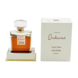 May include: A vintage perfume bottle of "Indiscret" by Lucien Lelong, presented in a white box. The perfume bottle is a square shape with a clear stopper and a gold-colored liquid inside. The box has the perfume name and brand.