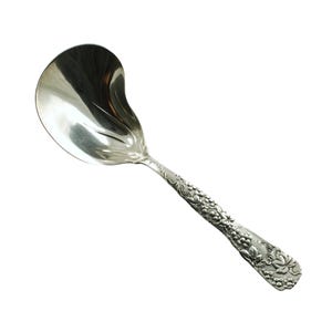 May include: A silver serving spoon with a curved bowl and a handle decorated with floral and grape designs. The spoon is made of a shiny, reflective metal and is designed for serving food. The spoon is approximately 25 cm long.