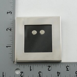 May include: A square, silver-toned picture frame with a black interior and two small, round, silver-toned objects. The frame is approximately 3 inches wide.