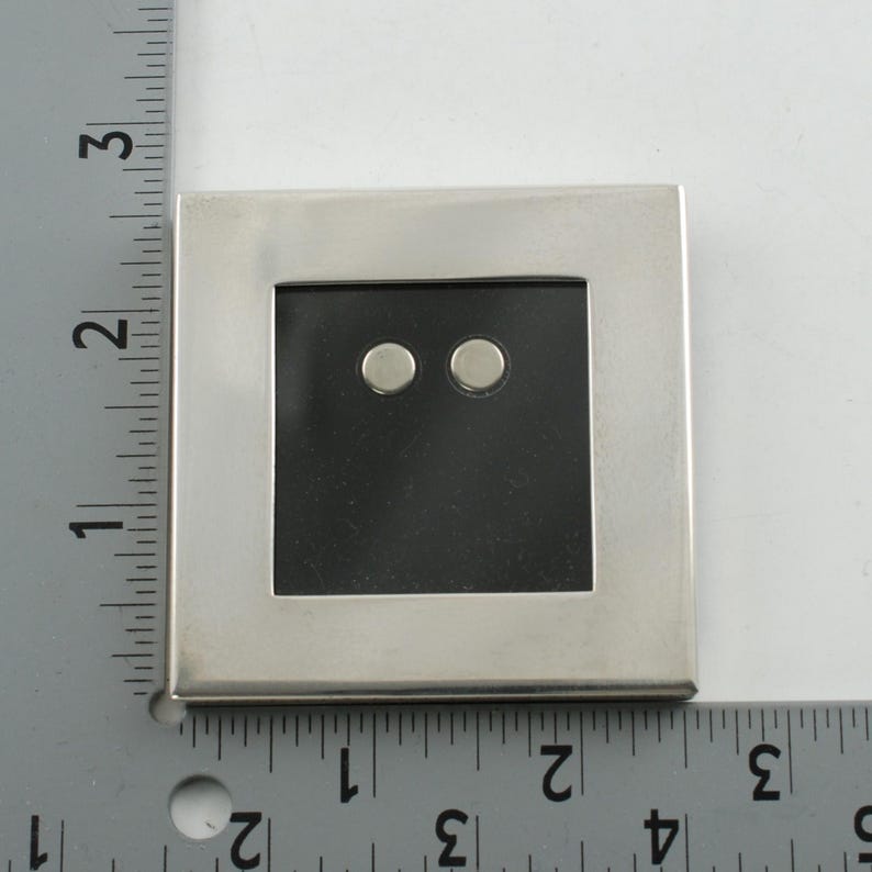 May include: A square, silver-toned picture frame with a black interior and two small, round, silver-toned objects. The frame is approximately 3 inches wide.
