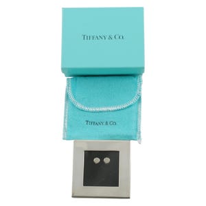 May include: A silver-toned square jewelry display with two small, round studs. The display sits below a light blue pouch and a matching box, both bearing the text "TIFFANY & Co.". The box is open, revealing the contents.