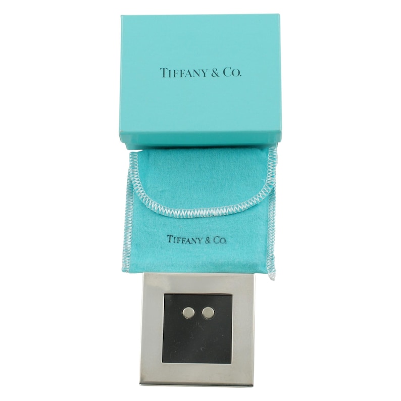 May include: A silver-toned square jewelry display with two small, round studs. The display sits below a light blue pouch and a matching box, both bearing the text "TIFFANY & Co.". The box is open, revealing the contents.