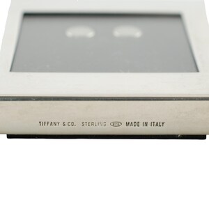 May include: A rectangular, silver-toned jewelry display case. The text "TIFFANY & CO. STERLING 925 MADE IN ITALY" is printed on the front. The case has a reflective, gray interior.
