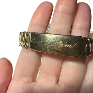 May include: A gold-toned bracelet with the inscription "Daughter Alexis" in cursive script. The bracelet features a rectangular plaque with decorative links on either side. The bracelet is held in a hand.