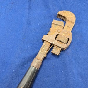 May include: An antique rusty pipe wrench with a black wooden handle. The wrench has a toothed jaw and an adjustable mechanism. The tool is designed for gripping and turning pipes or other cylindrical objects. The wrench is a vintage tool.