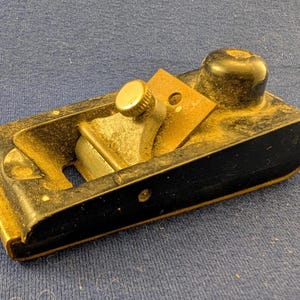 May include: A well-worn hand plane, with a black body and gold-coloured trim, and a brass adjustment knob. The tool shows signs of use, indicating it's a woodworking tool. The blade is visible within the plane's body.