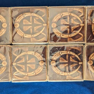 May include: Eight square decorative tiles with a brown and gold design. Each tile features a central oval shape with a cross-like pattern and a circular accent. The tiles are wrapped in clear plastic.