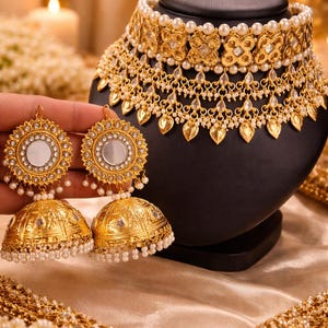 May include: Gold-toned jewelry set featuring a necklace and earrings. The necklace has a pearl-studded choker and ornate pendant. The earrings are round with mirrored centers and dangling pearl accents. The set is displayed on a black bust.