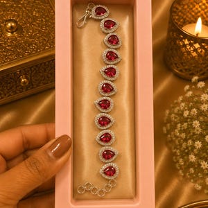 May include: A silver-toned bracelet featuring a series of eleven pear-shaped, deep red stones, each surrounded by a halo of small, clear, sparkling stones. The bracelet includes a lobster claw clasp and an adjustable chain link extension at the end.