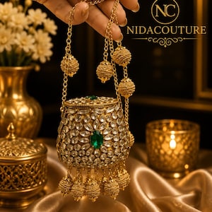 May include: A gold-toned potli bag featuring an intricate surface covered in clear, faceted stones and a central oval green stone. The bag has a gold chain handle with hanging gold bead charms and a circular lid, branded with the text NIDACOUTURE.