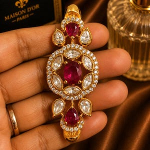 May include: A gold-toned pendant featuring a central oval-cut deep red gemstone surrounded by marquise-cut clear stones and small round pavé-set crystals. The design includes two additional pear-cut red gemstones at the top and bottom ends.